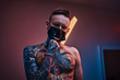 © Fxquadro - Bold and rebellious male model posing in a neon studio wearing a medical mask on his face, yellow sunglasses and tattooed in a japanese irezumi style on a half-naked body, thinking pose