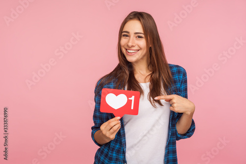 Fotomural I like this content! Charming positive girl in checkered shirt smiling playfully and holding heart icon, social media button to enjoy trendy interesting blog