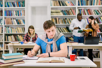 Concentrated handsome guy studying and writing notes from the book, sitting at the table in university library, preparing for exams, while his colleagues reading books and discussing on the background