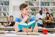 © sofiko14 - Attractive thinking handsome male student sitting at the table with many books and preparing for test or exam. Multiethnical students working together and library interior on the background