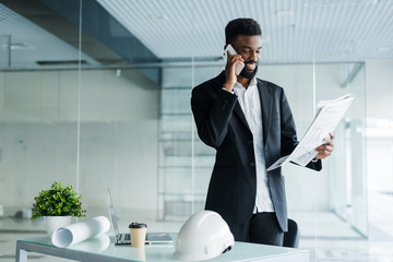 Smiling african american businessman talking on the phone with newspaper and cup of coffee in office