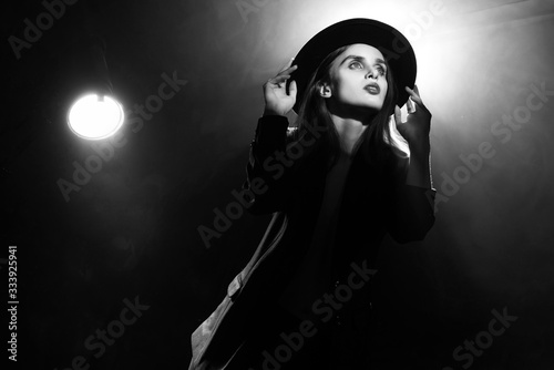 Cuadros en Lienzo Beautiful braless actress girl, wearing an unbuttoned blazer and a hat, poses and dances in the warm light rays of scenic illuminators in the theatrical fog