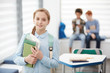 © Seventyfour - Waist up portrait of cute blonde schoolgirl holding books and smiling at camera while standing in school classroom, copy space