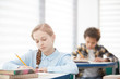 © Seventyfour - Portrait of cute blonde girl writing test while sitting at desk in school classroom, copy space