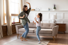 Full Length Overjoyed Barefoot Young Mommy Babysitter Dancing To Energetic Music With School Aged Girl On Floor Carpet At Home. Excited Mother Having Fun With Small Child Daughter In Living Room.