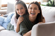 Overjoyed young attractive mother holding smiling sincere daughter on back, lying together on cozy sofa in living room. Happy two generations family looking at camera, enjoying tender moment at home.