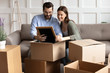 © fizkes - Full length happy young family couple unpacking belongings, looking at family photo in frame. Smiling spouses unboxing decorations, sitting together on comfortable couch between cardboard boxes.