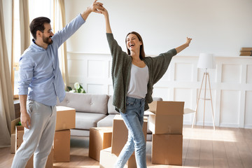 Happy young bearded man holding hand, twisting joyful wife in new living room near carton boxes. Excited married couple celebrating moving day in apartment house, dancing to energetic music.