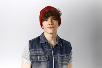 Wall Mural - Studio shot of handsome young twenty year old man posing against blank gray background with copy space for information wearing blue denim vest and red hat, having confident facial expression