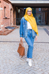 Wall Mural - Beautiful Arabic muslim student woman wearing yellow hijab, stylish female portrait over city street.