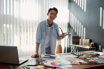 Wall Mural - happy at work. cheerful asian man holding eyeglasses and smiling at camera while leaning on a table 