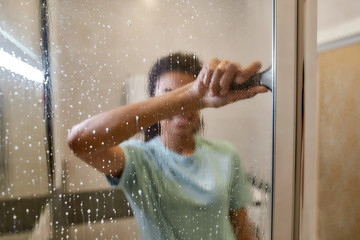 Making your home sparkling clean. Young smiling afro american woman in uniform cleaning shower door with squeegee and spray detergent