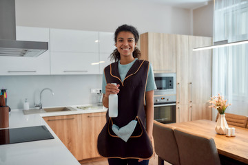 Wall Mural - Professional Service. Young cheerful afro american woman holding spray detergent, looking at camera and smiling while cleaning in the kitchen. Professional female cleaner in uniform