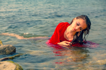 Young long-haired brunette in red beach dress reclining in the turquoise water of the ocean on a hot day. Beautiful sad woman posing in the sea during vacation.