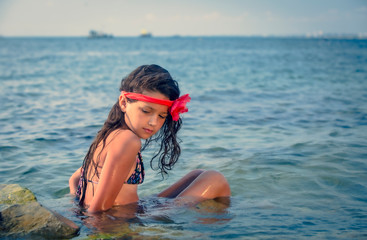 Mysterious dreamy young girl with a red bandage on her head sitting in the water on a rocky beach