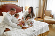© Svitlana - Enjoy unforgettable experience. Parents and two kids in white bathrobes smiling while having breakfast in bed, eating and drinking in luxurious hotel room. Family, resort, room service concept