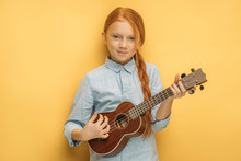 Portrait Of Awesome Caucasian Child Girl Playing Ukulele, Attractive Girl With Natural Red Hair Keen On Music And Instruments. Isolated Yellow Background