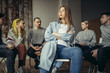 © Roman - young caucasian alcoholic woman tired of conviction and gossip, sit in the center of room while people in the background discuss problems connected with alcohol. support, problems, help, desperate
