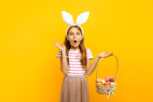 A Shocked Surprised Girl On A Head With Rabbit Ears, Holding A Wicker Basket With Colored Eggs On A Yellow Background