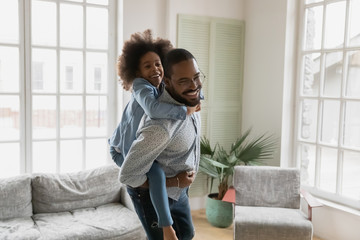 Wall Mural - African ethnicity father piggy back riding her little daughter having fun together in cozy light modern living room. Cheerful kid girl hanging on a back of daddy laughing enjoy active weekend at home