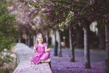 Wall Mural - Beautiful girl 10 y.o. in pink dress sit in cherry blossom park on a spring day, flower petals falling from the tree