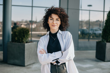 Charming caucasian woman with curly hair and glasses is posing with crossed hands and looking at camera