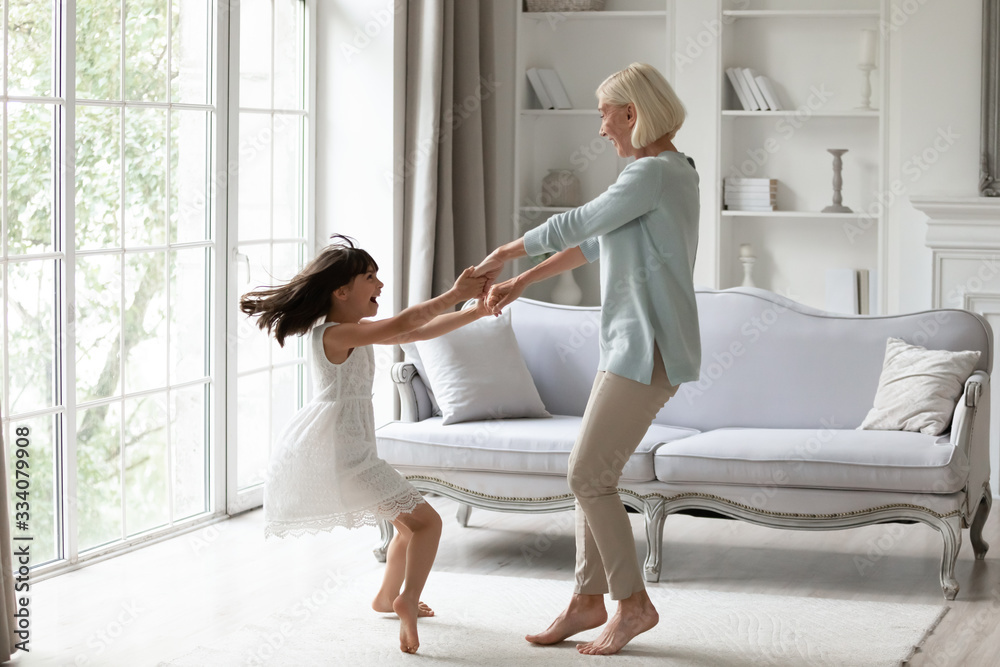 Overjoyed senior grandmother dancing entertaining with little granddaughter in living room, happy grandparent have fun engaged in funny family activity with small grandchild at home together - obrazy, fototapety, plakaty 