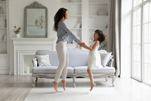 Happy Young Mom And Cute Little Preschooler Daughter Have Fun Dancing Jumping In Living Room Together, Overjoyed Millennial Mother Or Nanny Feel Playful Entertain At Home With Small Girl Child