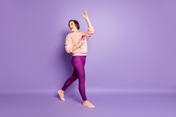 Wall Mural - Full length profile photo of pretty lady holding telephone walking down street waving palm greeting friends wear casual stylish pink sweater trousers isolated purple color background