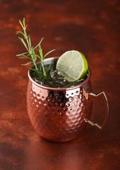 Moscow mule cocktail in a copper mug with lime and rosemary on brown background