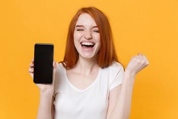 Wall Mural - Joyful young redhead woman girl in white t-shirt posing isolated on yellow background. People lifestyle concept. Mock up copy space. Hold mobile phone with blank empty screen, doing winner gesture.