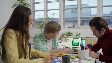 Wall Mural - Group of three young marketing specialists and designers sitting at desk and discussing website interface templates for various devices presented on laptop screen