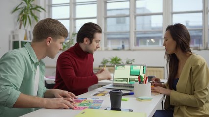 Wall Mural - Zoom out and in shot of team of three marketing specialists and designers sitting at desk in office and discussing website templates for different devices presented on laptop and printed on paper