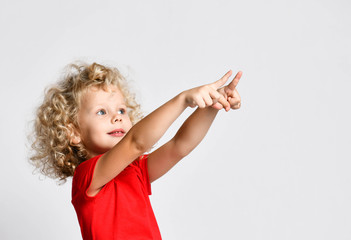 Curly hair blonde kid girl in red t-shirt holds pointing fingers at free copy space at upper corner