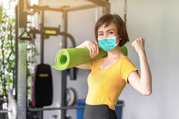 Young woman in sportswear doing work-out, sports training, gymnastics and wearing mouth mask against the corona virus. She's holding a training mat. Home fitness.