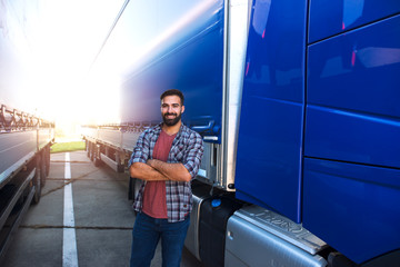 Wall Mural - Portrait of young Caucasian bearded trucker with arms crossed standing by his truck vehicle. Transportation service.