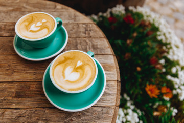 Light blue cups of hot fresh coffee with heart-shaped drawing made of milk foam on wooden table with blur background of flower bush. Aroma drinks made by professional barista for two persons.