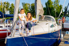 Portrait Of Smiling Mother And Daughter On Prow Of Sailboat Or Yacht Anchored In Marina At Bright Sunny Day. Blurred Background With Boats And Trees