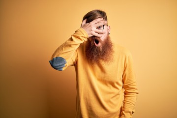 Sticker - Handsome Irish redhead man with beard wearing glasses over yellow isolated background peeking in shock covering face and eyes with hand, looking through fingers with embarrassed expression.