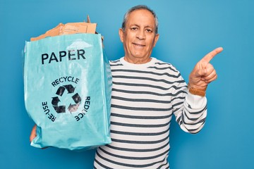 Wall Mural - Senior grey-haired man recycling holding bag with cardboard to recycle over blue background very happy pointing with hand and finger to the side