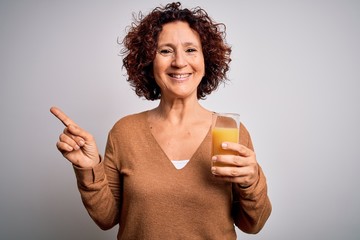 Poster - Middle age curly hair woman drinking healthy glass of orange juice over white background very happy pointing with hand and finger to the side