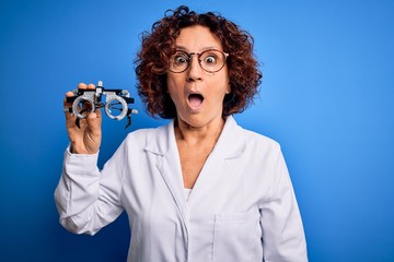 Poster - Middle age optical woman wearing coat holding optometry glasses over blue background scared in shock with a surprise face, afraid and excited with fear expression