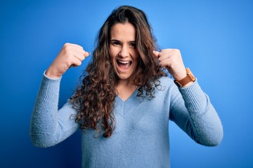 Poster - Young beautiful woman with curly hair wearing blue casual sweater over isolated background angry and mad raising fists frustrated and furious while shouting with anger. Rage and aggressive concept.