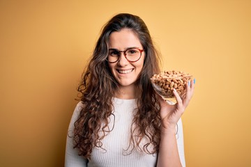 Sticker - Beautiful woman with curly hair holding bowl with healthy peanuts over yellow background with a happy face standing and smiling with a confident smile showing teeth