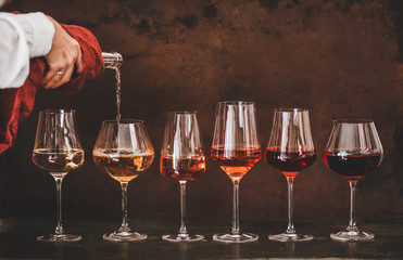 Shades of Rose wine in stemmed glasses placed in line from light to dark colour and womans hand pouring wine from bottle to glass, rusty brown background behind. Wine bar, wine shop, tasting concept