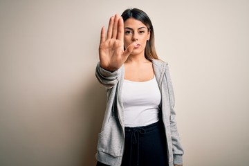 Poster - Young beautiful brunette sportswoman wearing sportswoman training over white background doing stop sing with palm of the hand. Warning expression with negative and serious gesture on the face.