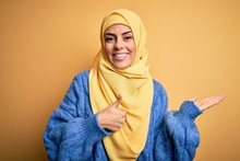 Young Beautiful Brunette Muslim Woman Wearing Arab Hijab Over Isolated Yellow Background Showing Palm Hand And Doing Ok Gesture With Thumbs Up, Smiling Happy And Cheerful
