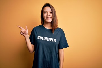 Young beautiful brunette girl doing volunteering wearing t-shirt with volunteer message word cheerful with a smile on face pointing with hand and finger up to the side with happy and natural