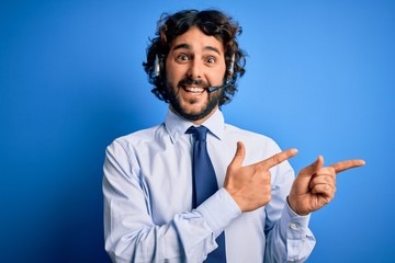 Young handsome call center agent man with beard working using headset over blue background smiling and looking at the camera pointing with two hands and fingers to the side.