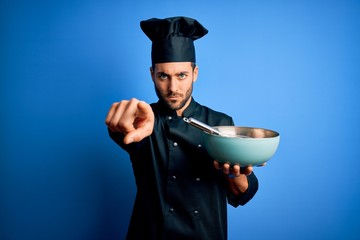 Canvas Print - Young cooker man with beard wearing uniform using whisk and bowl over blue background pointing with finger to the camera and to you, hand sign, positive and confident gesture from the front